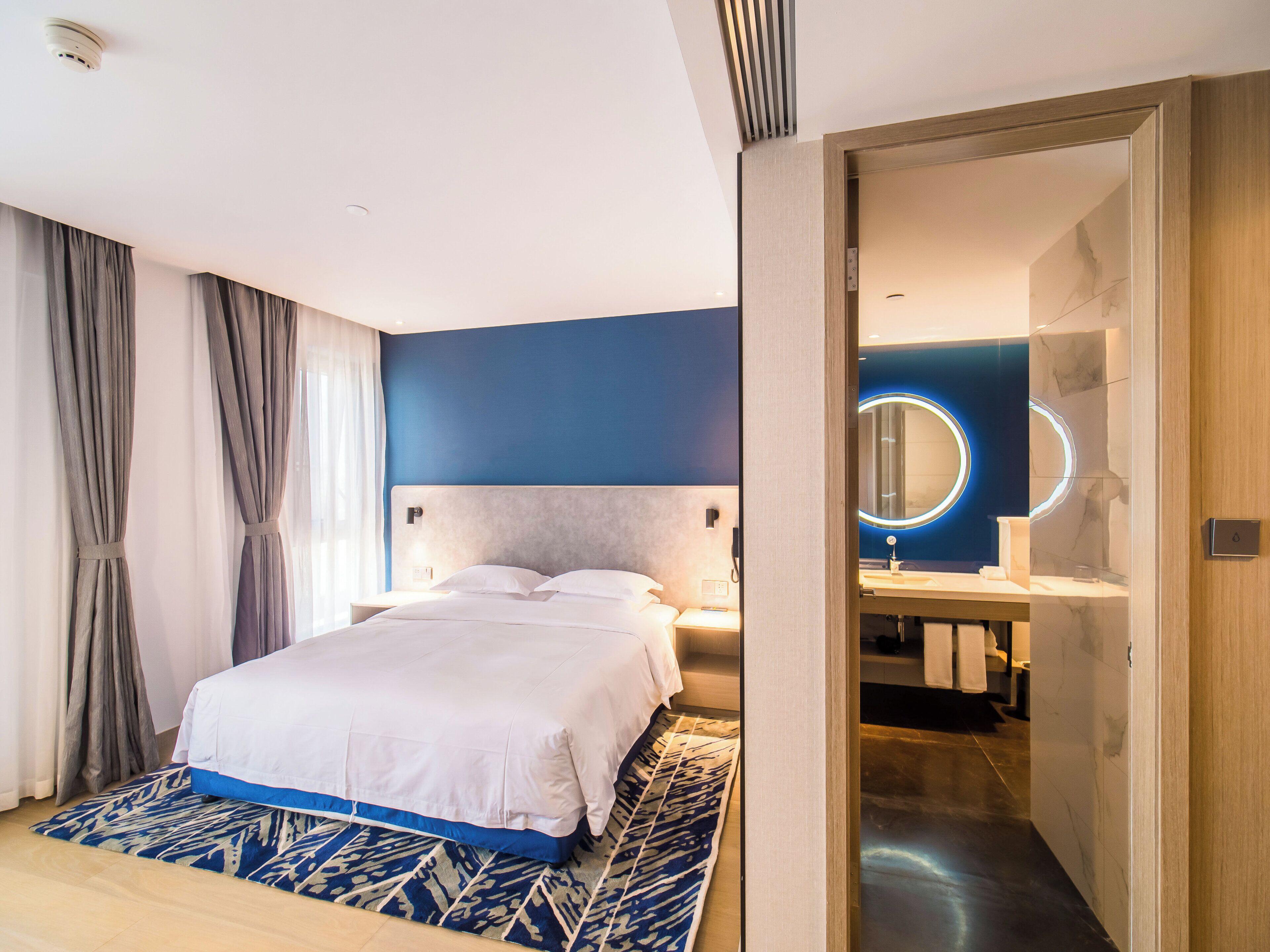 A modern hotel room with a bed, bedside tables, a bathroom with a mirror, and a patterned rug.
