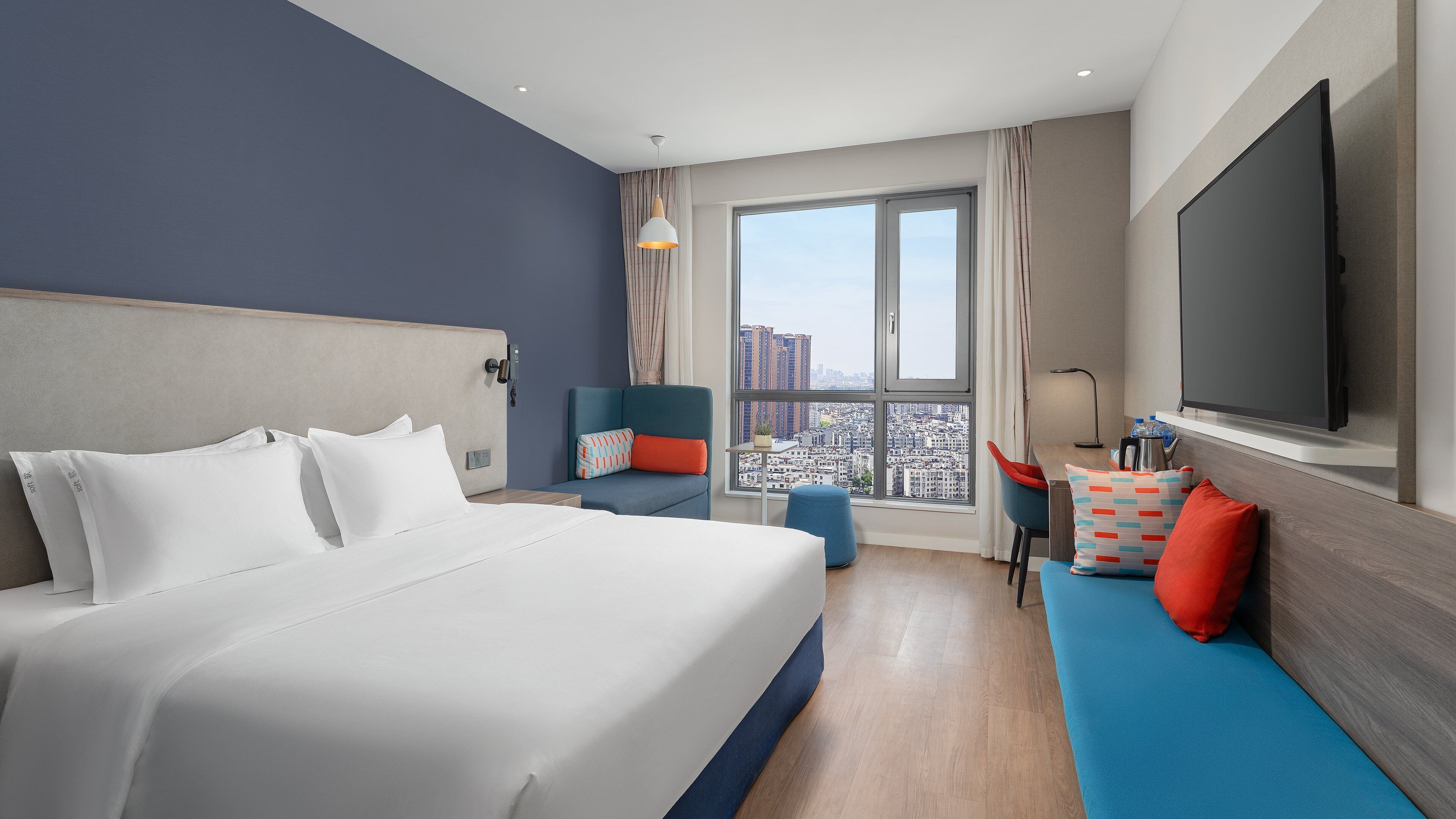 A modern hotel room with a large bed, a seating area with a TV, and a city view through the window.