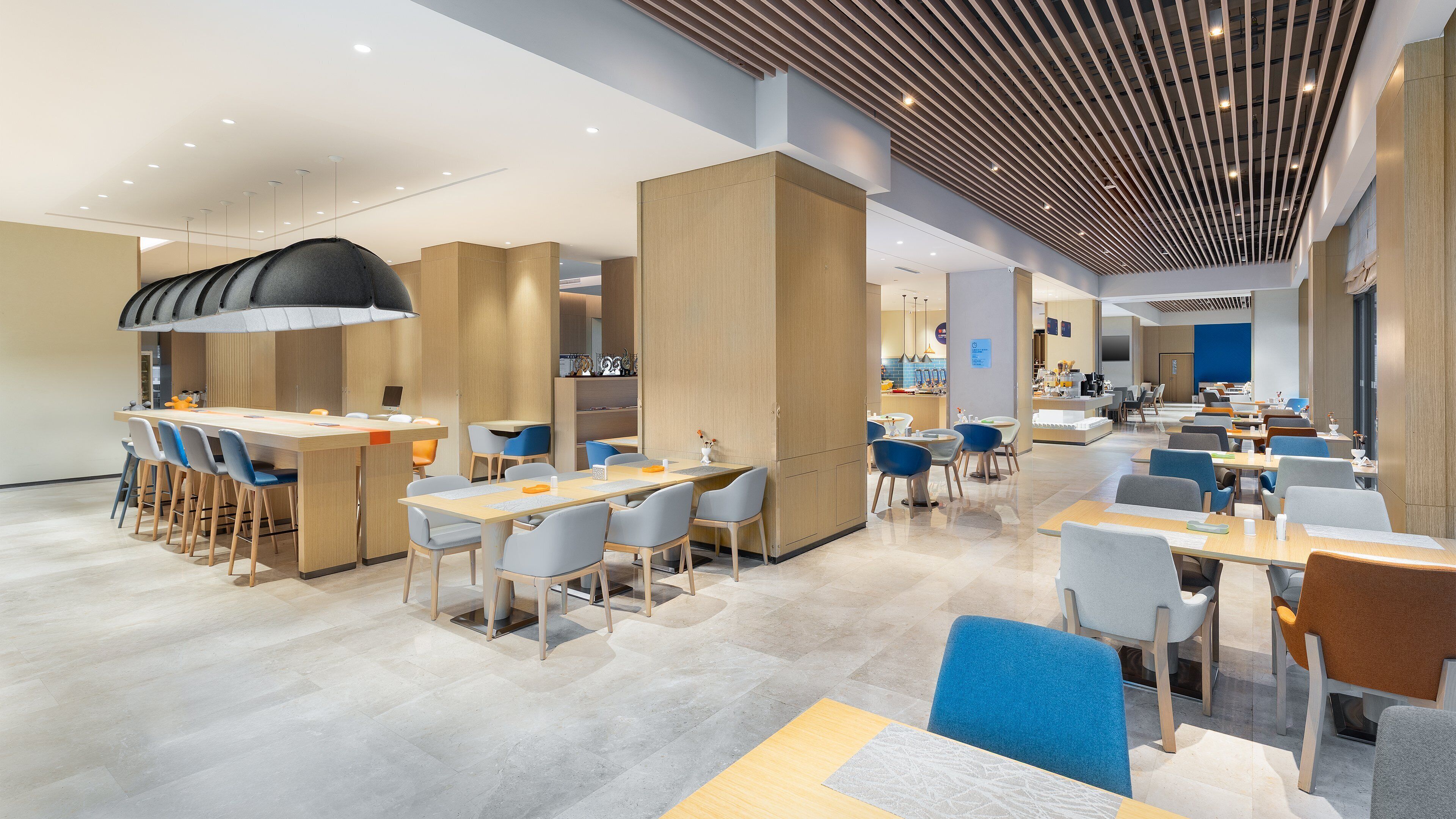 A modern, spacious dining area with multiple tables and chairs, a central bar area, and a high ceiling with linear lighting.