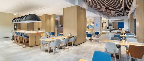 Un espace de restauration moderne et spacieux, doté de nombreuses tables et chaises, d’un bar central et d’un plafond haut avec un éclairage linéaire.