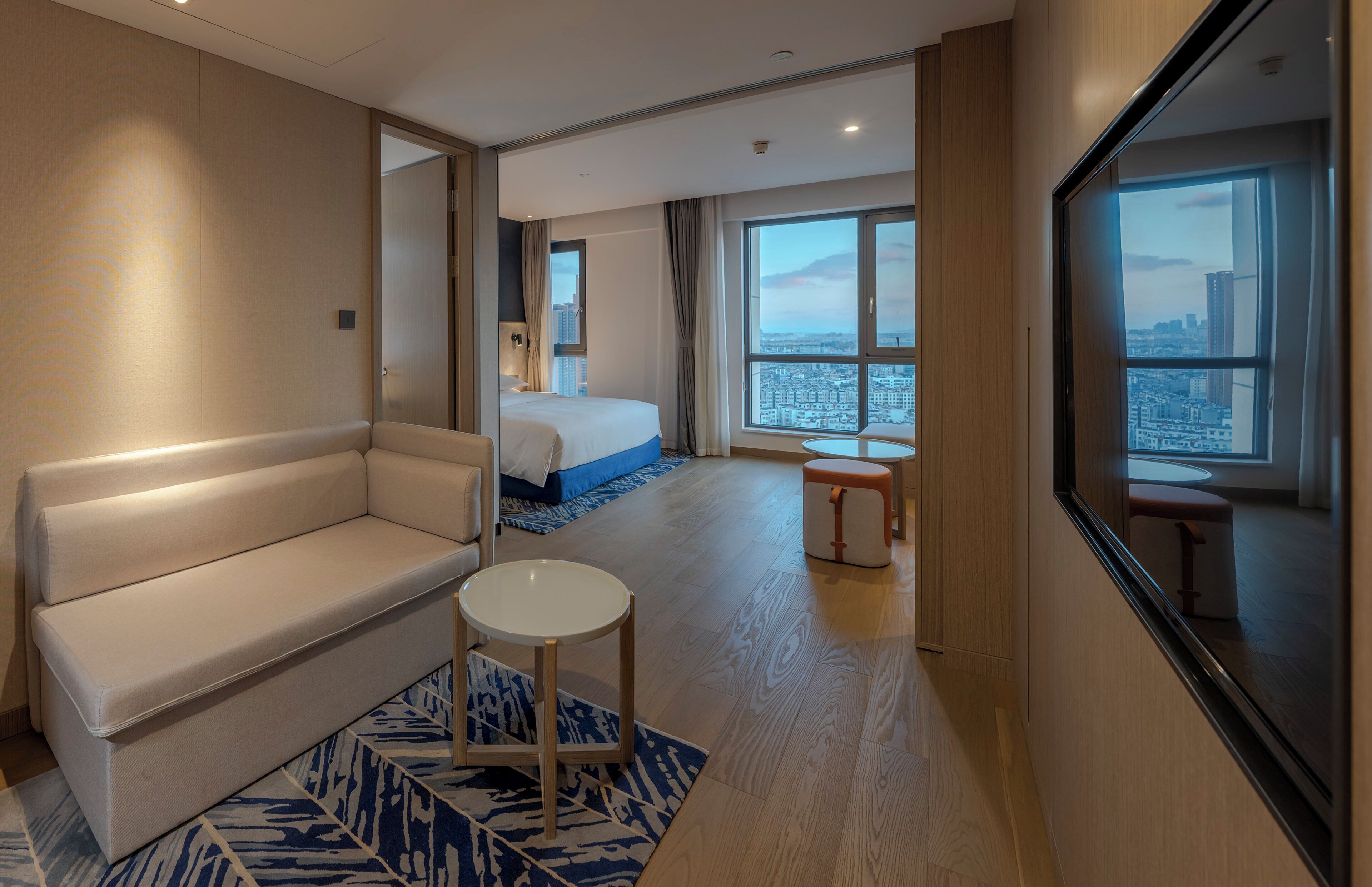 A hotel room with a sofa, a small round table, a bed with a blue accent, a large window with a city view, and a mirror on the wall.