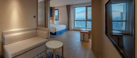 Habitación de hotel con sofá, una mesita redonda, cama con detalles azules, ventana grande con vista a la ciudad y espejo en la pared.