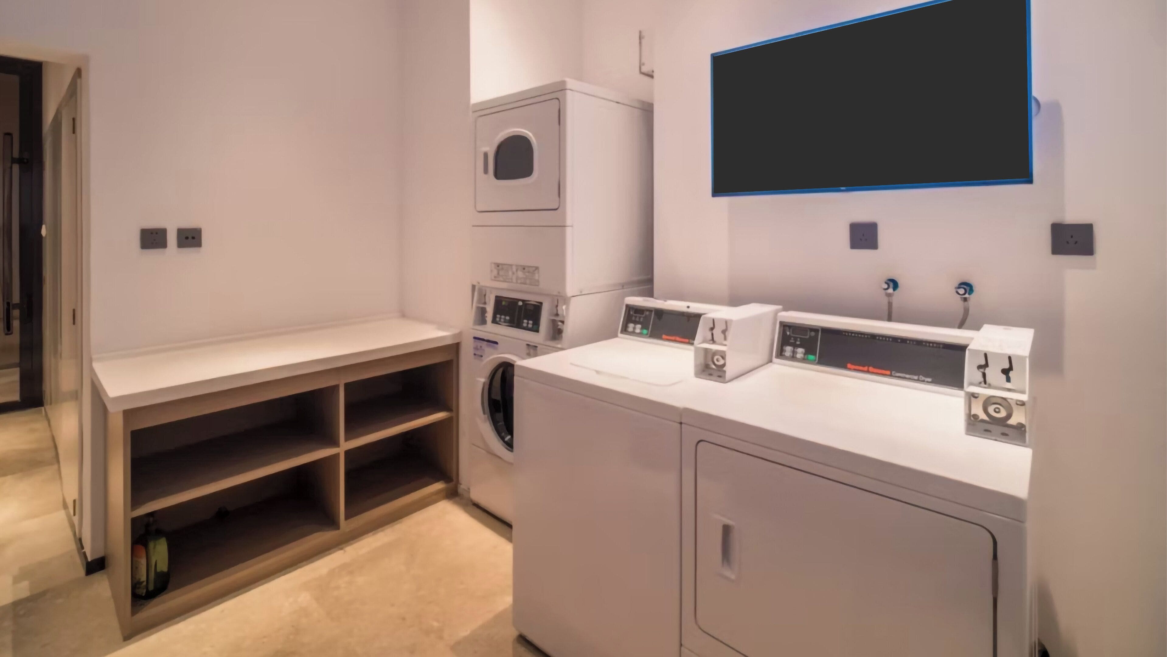 A laundry room with a washing machine, dryer, and stacked dryers. There is a bench and a shelf with a bottle.