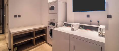 A laundry room with a washing machine, dryer, and stacked dryers. There is a bench and a shelf with a bottle.