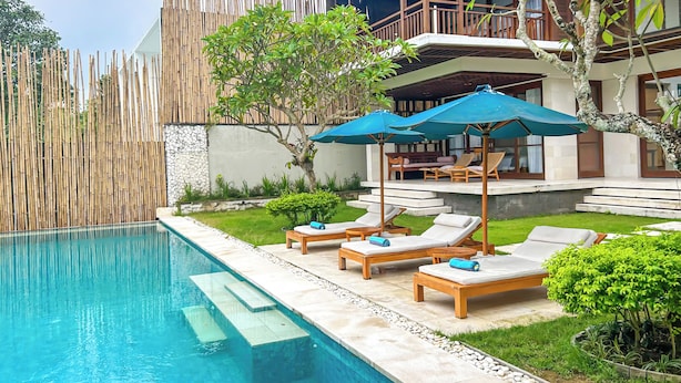 A poolside area with lounge chairs and umbrellas, a wooden fence, and a building with a balcony. - Villa Rusa Biru - Peaceful Ricefield Views in Central Canggu (Kecamatan Kuta Utara)