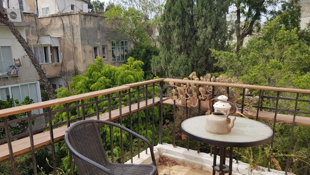 A balcony with two chairs and a table, a decorative plant, and a view of a building and trees. - Specious room next to the beach (Tel Aviv-Yafo)