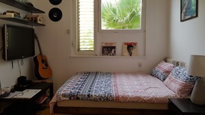 A bedroom with a bed, a guitar, a TV, and a window with blinds. - Specious room next to the beach (Tel Aviv-Yafo)