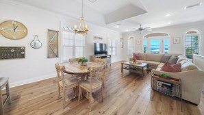 A spacious living room with a wooden floor, a dining area with a round table and chairs, a sofa, and a TV. - Pier Park only 50 Steps to the beach! Pet friendly. Ocean View.  (Panama City Beach)