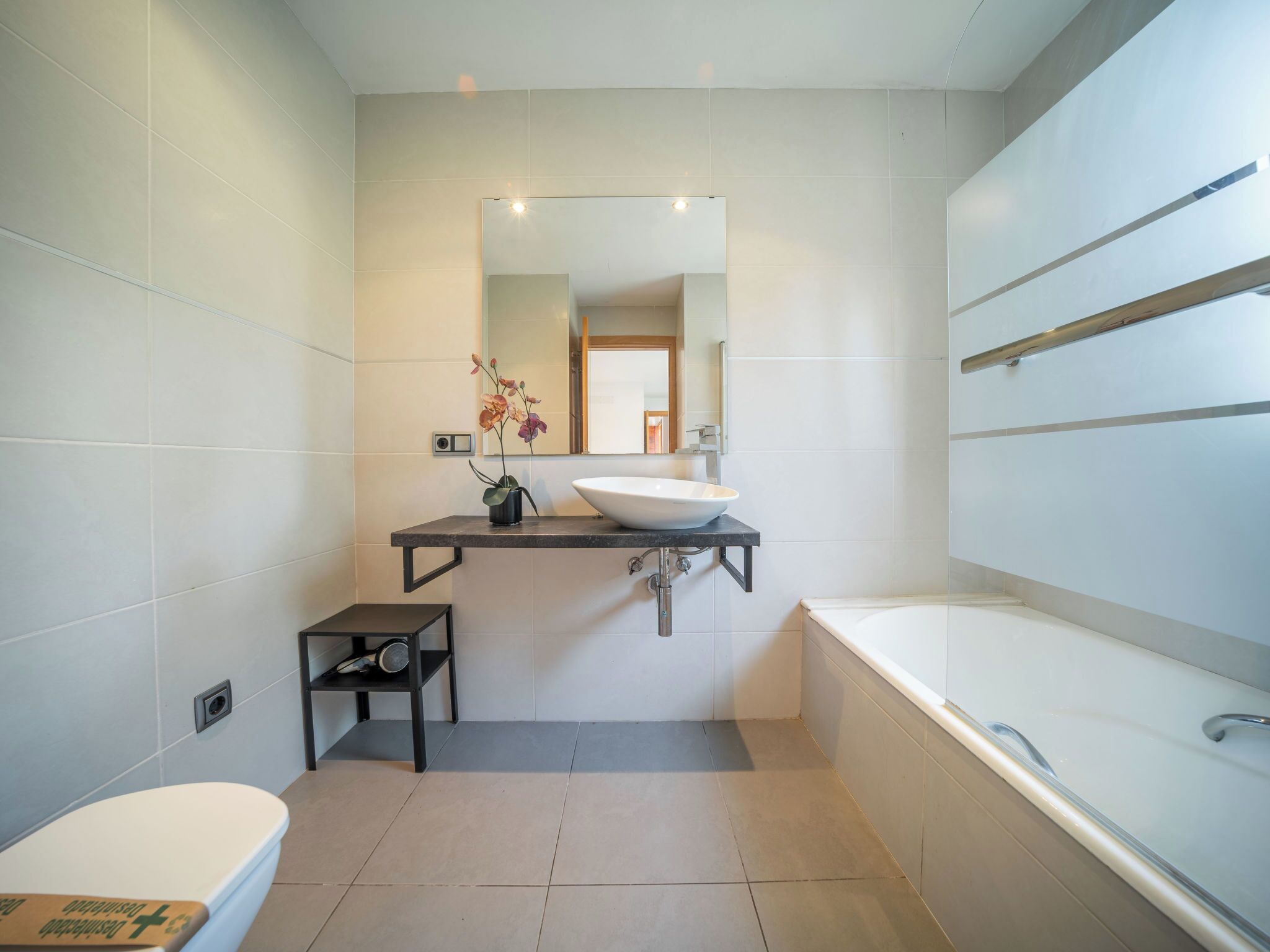 Een moderne badkamer met een wit wastafelmeubel, een grote spiegel, een klein krukje en een bad.
