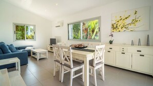 A modern living room with a sofa, a dining table with chairs, a TV, and a large window with a view of greenery. - Amanecer by Interhome (Alcúdia)