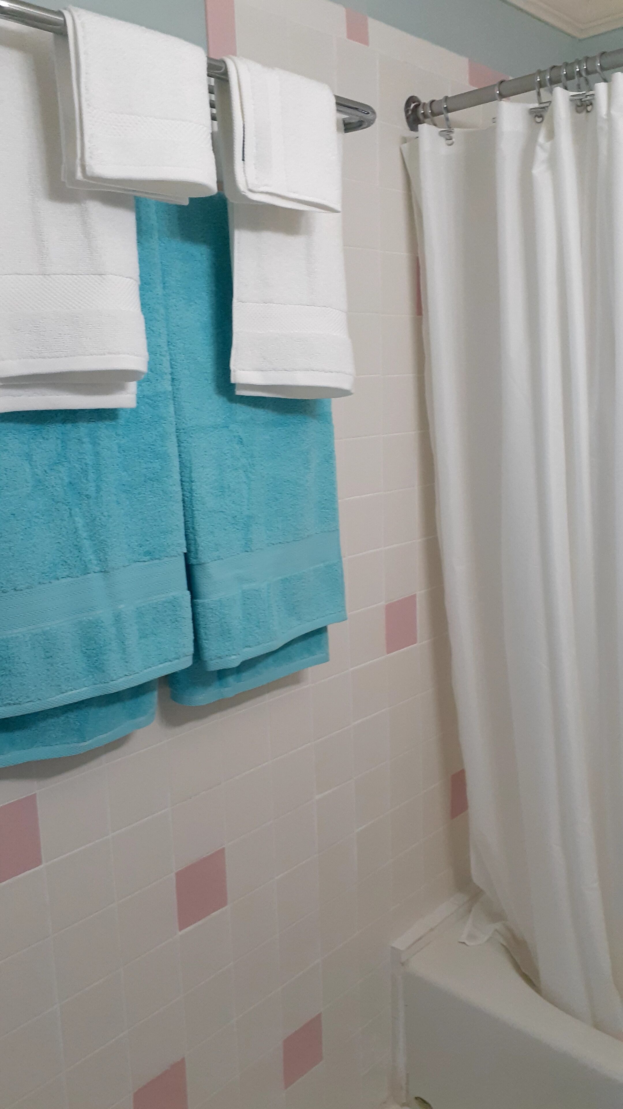 Une salle de bain avec deux serviettes blanches et une serviette bleue suspendues à un porte-serviettes, un rideau de douche blanc et un mur carrelé agrémenté de touches roses.