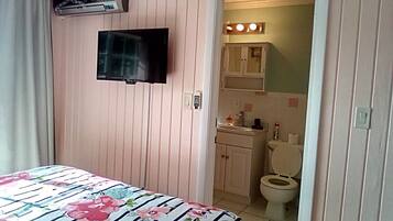 Et værelse med en seng, et fjernsyn ophængt på væggen, et badeværelse med toilet og vask, og en dør der fører til et andet værelse.
