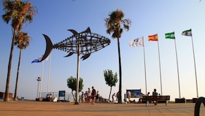 A public square with palm trees, a large metal sculpture, and flags.