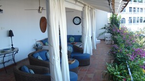 A covered outdoor seating area with wicker furniture and blue cushions, a clock on the wall, and a view of a building with balconies. - lovely suite getaway (Puerto Vallarta)