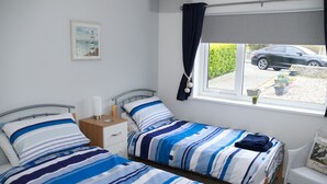 A bedroom with two beds, a window with curtains, a small table, and a chair. - Penmon View, Moelfre, Anglesey. With sea and mountain views. (Moelfre)