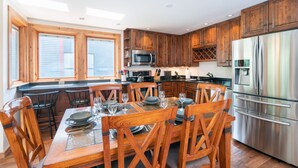 A modern kitchen with wooden cabinets, stainless steel appliances, and a dining area with wooden chairs and a glass table set for six. - Beautifully Remodeled - Steps to Lift 7 and River Trail with Mountain Views (Telluride)