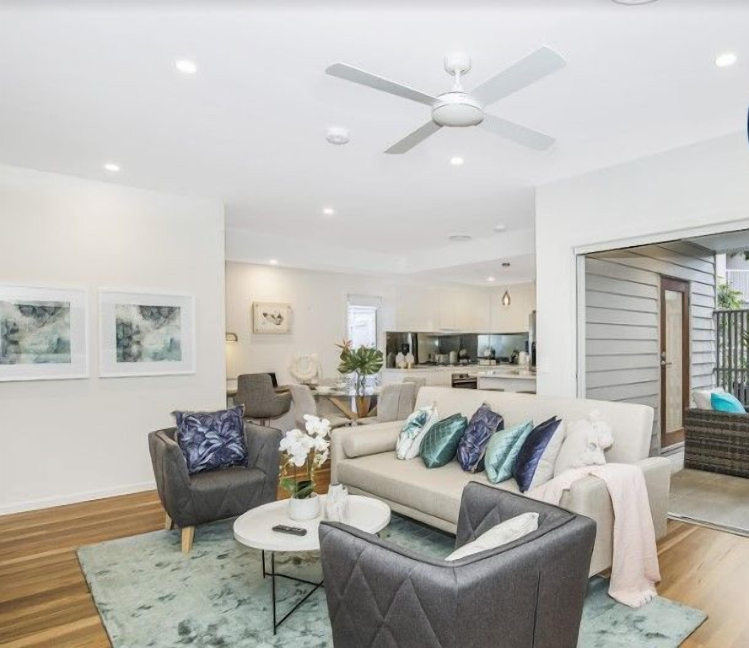 A modern living room with a sofa, armchairs, and a coffee table. There is a ceiling fan, a kitchen area in the background, and a sliding glass door leading to an outdoor patio.