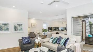 A modern living room with a sofa, armchairs, and a coffee table. There is a ceiling fan, a kitchen area in the background, and a sliding glass door leading to an outdoor patio.