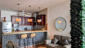Een moderne keuken met donkere houten kastjes, een roestvrijstalen koelkast en een achterwand met een patroon. In de woonkamer staan twee barkrukken en een grijze bank.