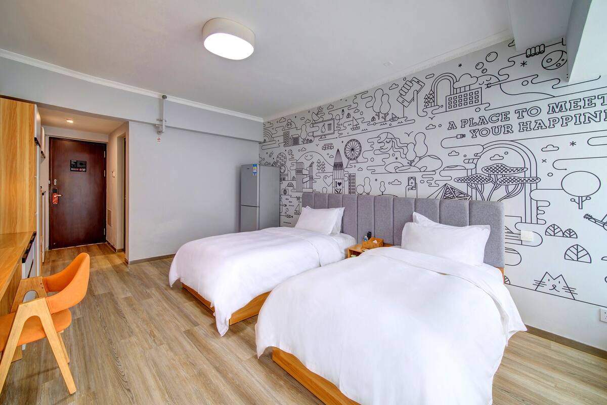 A hotel room with two beds, a desk, and a chair. There is a wall mural with various doodles.