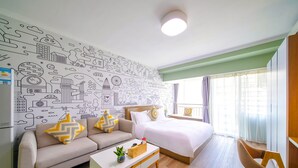 Quarto de hotel moderno com um mural de paisagem urbana, cama com roupa de cama branca, sofá bege com almofadas estampadas, mesa de centro de madeira e janela com cortinas.