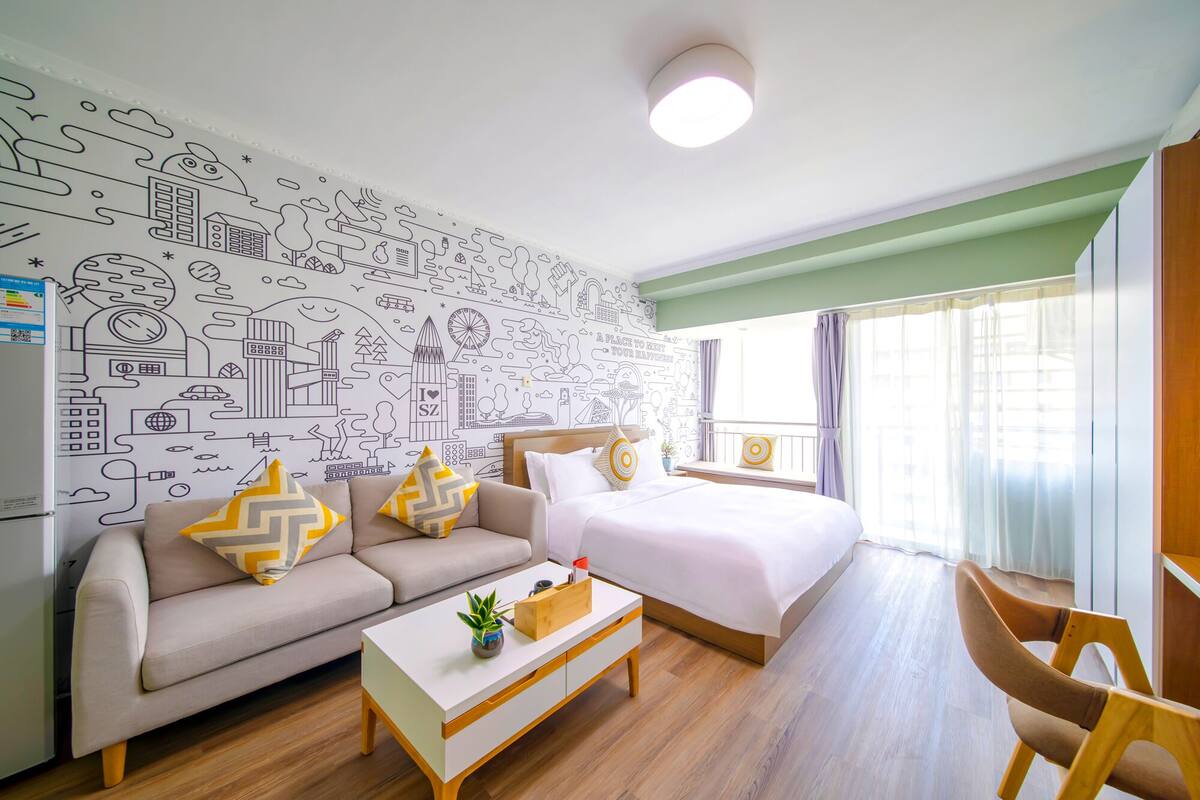 A modern hotel room with a cityscape mural, a bed with white bedding, a beige sofa with patterned cushions, a wooden coffee table, and a window with curtains.