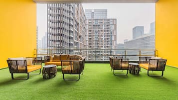 A room with a green artificial turf floor, a sofa, and two armchairs with cushions, facing a large window with a cityscape view.