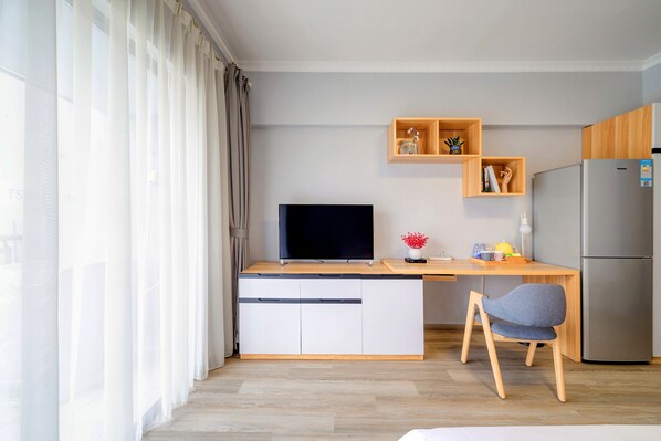 A compact living space with a kitchenette, a flat-screen TV, a wooden desk, a chair, and a large window with curtains. - Shenzhen T Hotel Apartment (Shenzhen)