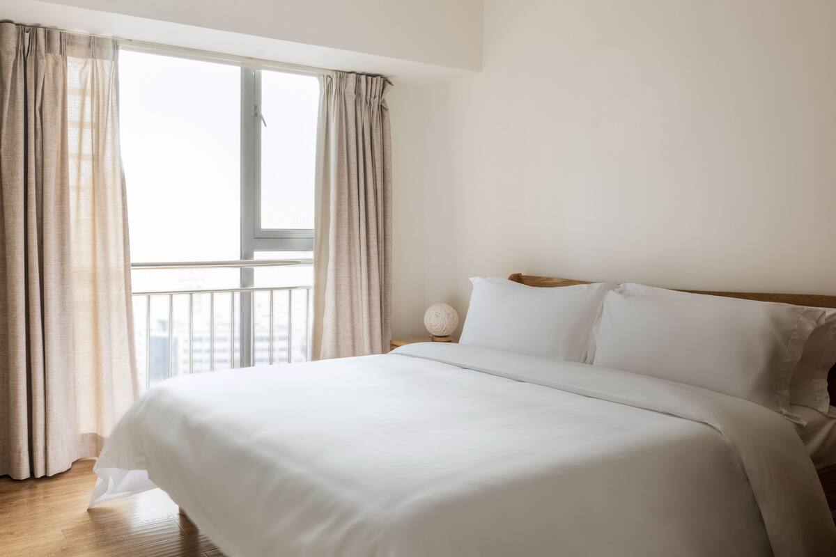 A hotel room with a large bed, a nightstand with a lamp, and a view of the city through a window with curtains.
