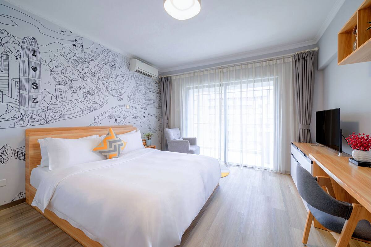 A hotel room with a large bed, a desk with a chair, a TV, and a large window with curtains.