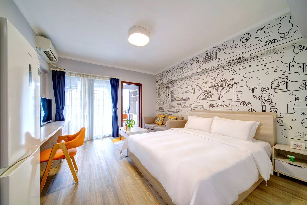 A modern hotel room with a large bed, a desk with a chair, a window with curtains, and a wall with travel-themed artwork.