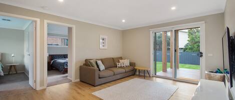 A modern living room with a sofa, a small table, and a rug. There is a door leading to a bedroom and a view of the outside.