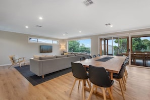 A modern living room with a large sofa, dining table, and chairs. There is a painting on the wall and a window providing natural light.