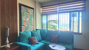 Un canapé de couleur turquoise agrémenté de coussins à motifs floraux, une petite table blanche et un tableau aux couleurs tropicales accroché au mur.