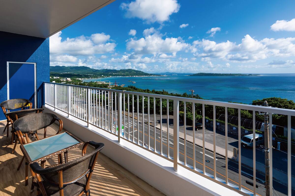 A balcony with wicker furniture, a glass table, and a view of a coastal area with mountains and a bridge.