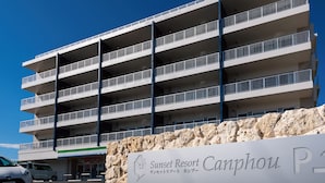 A multi-story building with balconies, a stone wall with a sign, and a parking area. - Sunset Resort Canphou (Motobu)