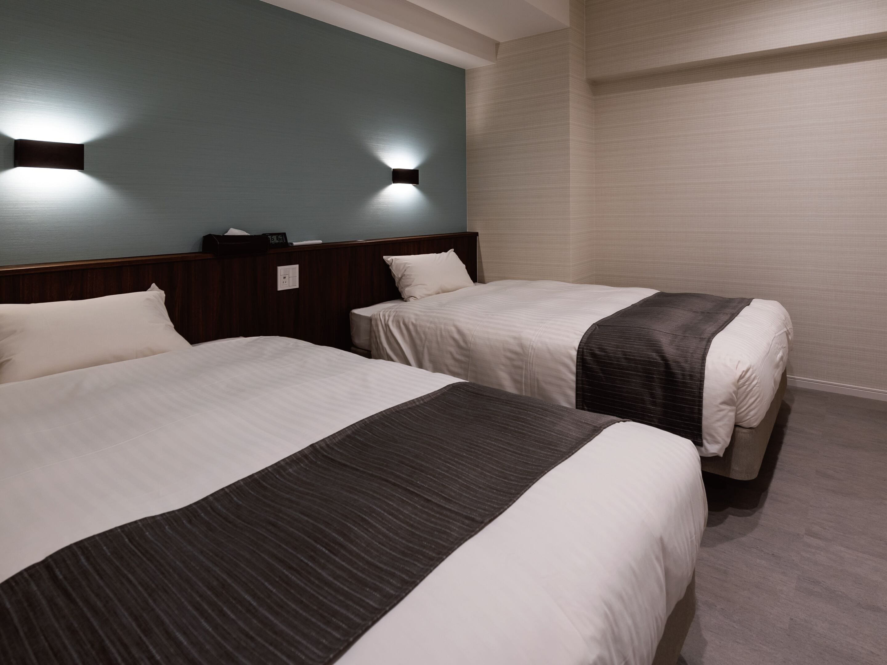 A hotel room with two beds, a wooden headboard, a wall-mounted light fixture, and a plain wall.