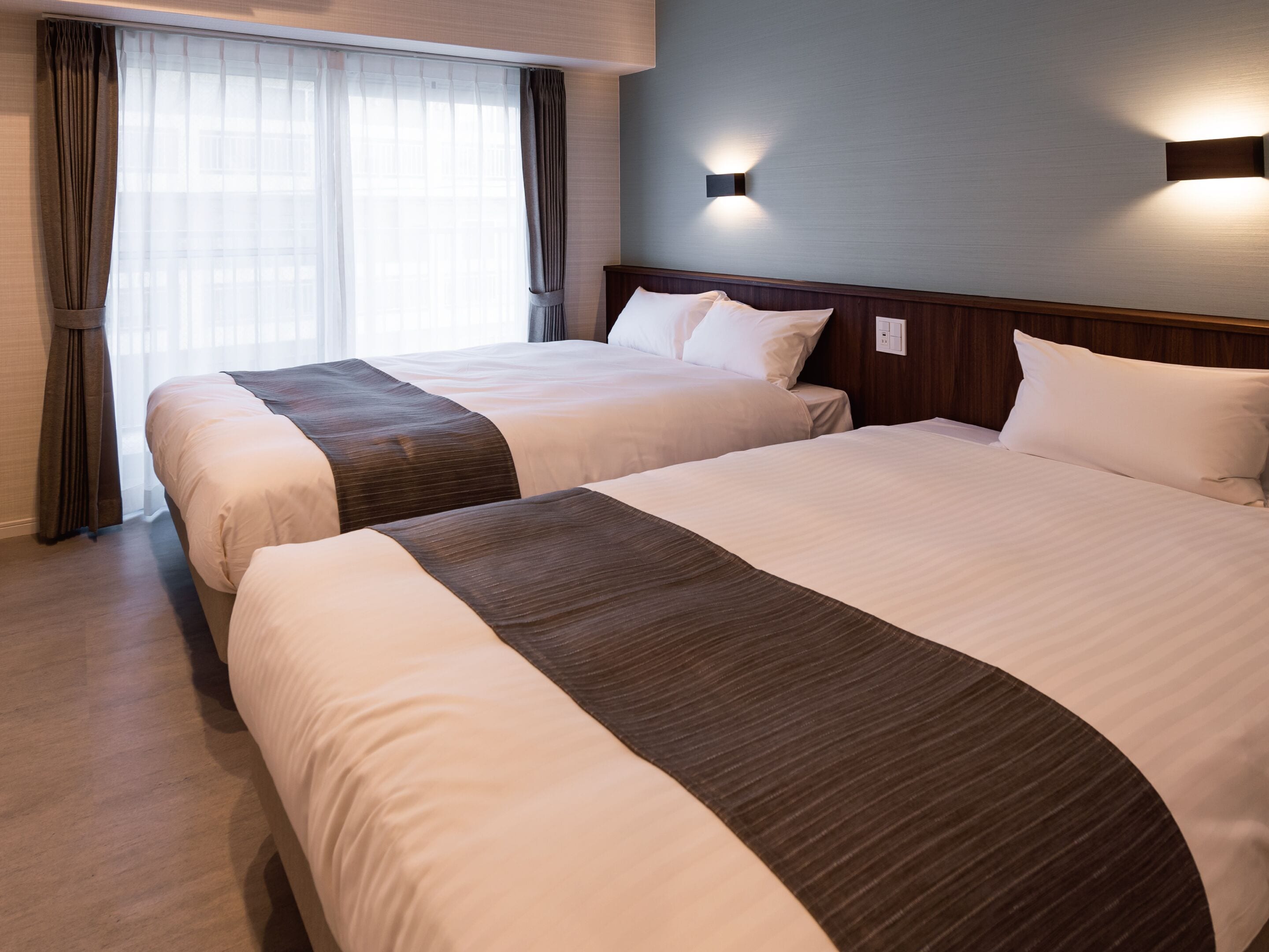 A hotel room with two beds, a wooden headboard, a wall-mounted light, and a window with curtains.