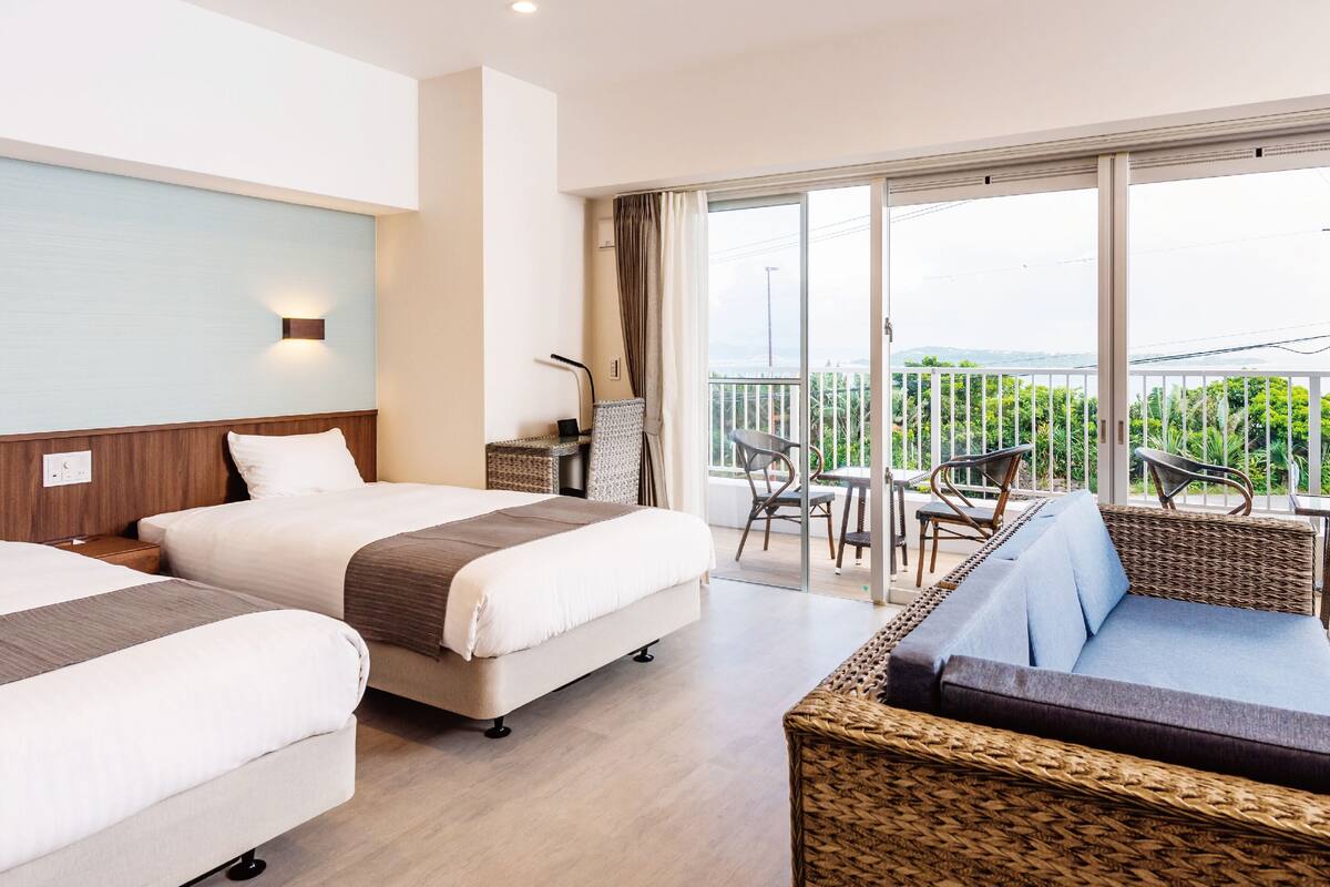 A hotel room with two beds, a balcony with wicker furniture, and a view of greenery.