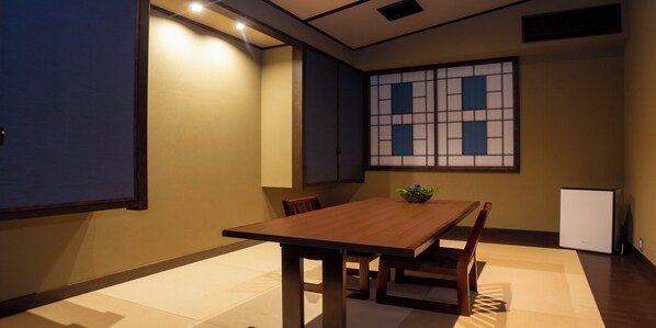 A room with a wooden dining table, chairs, a small white cabinet, and a traditional Japanese screen on the wall.