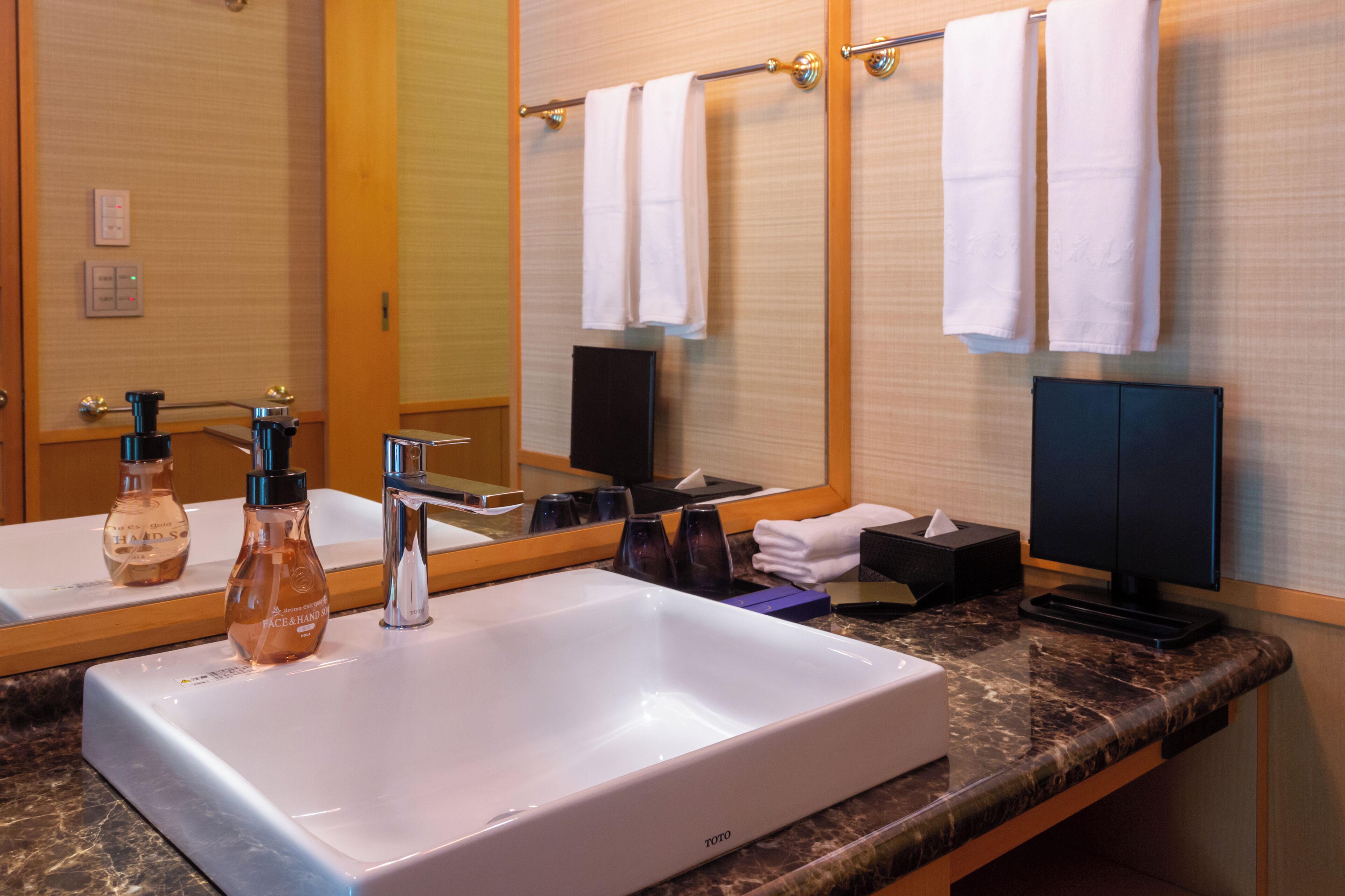 Un baño con un lavamanos grande de color blanco, dos botellas de jabón para manos, un espejo, un perchero con toallas blancas, un armario negro y un televisor negro.