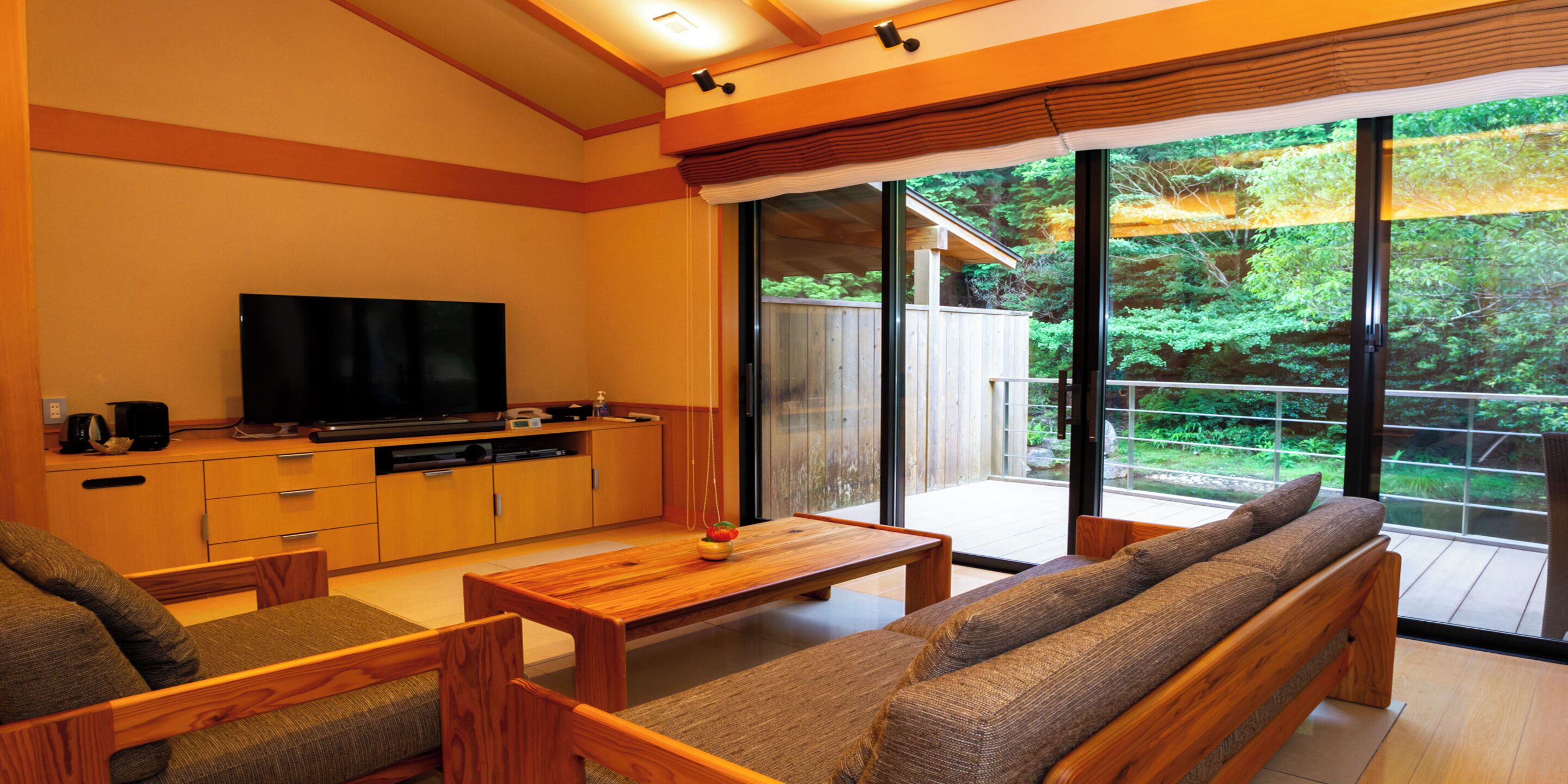 A modern living room with a flat-screen TV, wooden furniture, and a large sliding glass door leading to a balcony with a view of greenery.