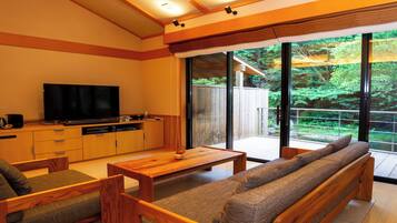 A modern living room with a flat-screen TV, wooden furniture, and a large sliding glass door leading to a balcony with a view of greenery.