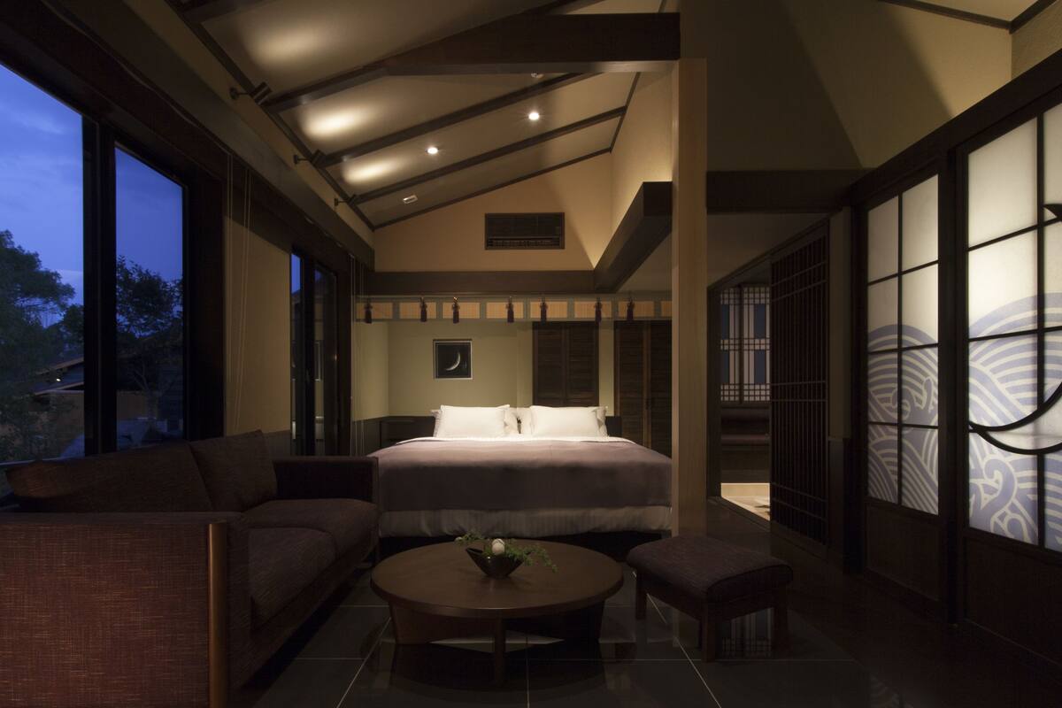 Habitación de hotel moderna con una cama grande, zona de estar con una mesa de centro y ventanas amplias que ofrecen vistas al exterior.