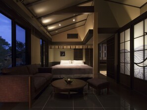 Habitación de hotel moderna con una cama grande, zona de estar con una mesa de centro y ventanas amplias que ofrecen vistas al exterior.