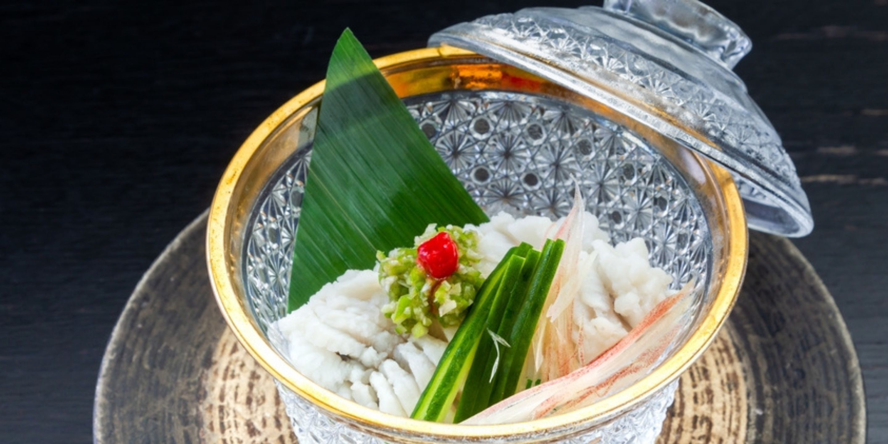 A glass bowl with a decorative lid, containing white rice, green vegetables, and a red fruit garnish.