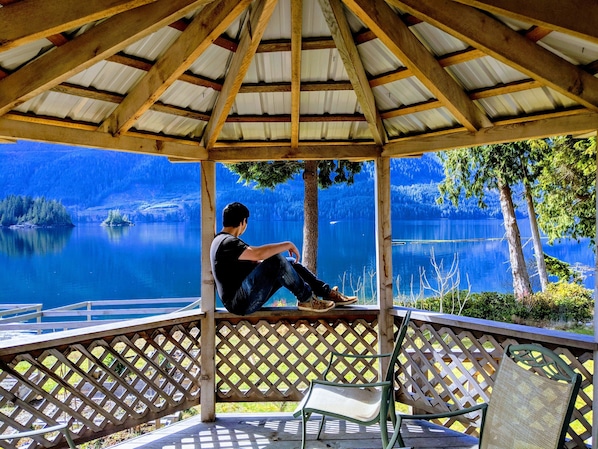 Un quiosco de madera con una persona sentada en un banco, contemplando un lago tranquilo con árboles y colinas al fondo.