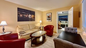 A living room with a sofa, two armchairs, a coffee table, and a TV. There are paintings on the walls and a door leading to another room.