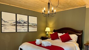 A neatly made bed with white and red bedding, a wooden headboard, and a chandelier above. There are three framed artworks on the wall.
