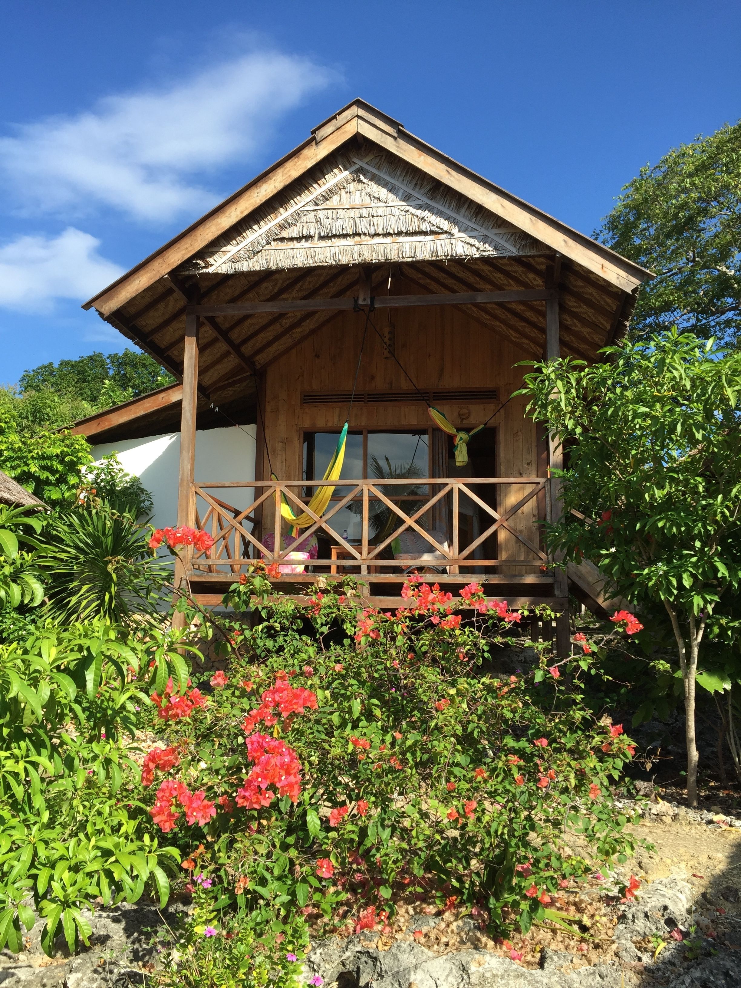A wooden house with a balcony, surrounded by lush greenery and flowering plants.